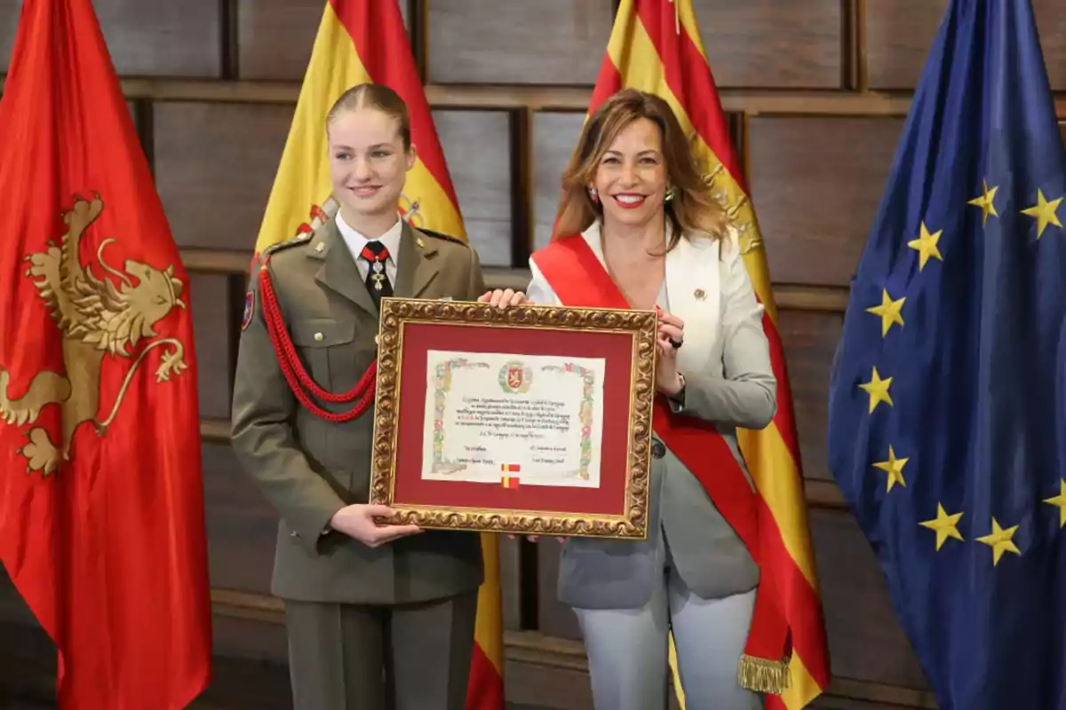 Dos mujeres posan con un diploma enmarcado, una de ellas vestida con uniforme militar y la otra con traje formal, frente a banderas de España y la Unión Europea.