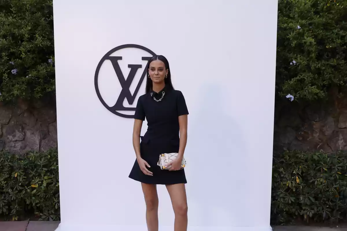 Una mujer posando frente a un fondo blanco con el logotipo de Louis Vuitton, vestida con un vestido negro y sosteniendo un bolso blanco.