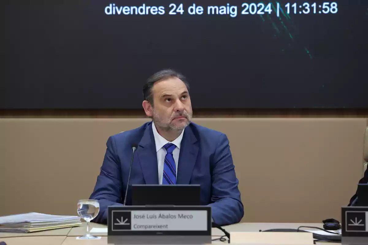 Un hombre con traje azul está sentado en una mesa con un micrófono y un vaso de agua frente a él, mirando hacia arriba, con una pantalla detrás que muestra una fecha y hora.