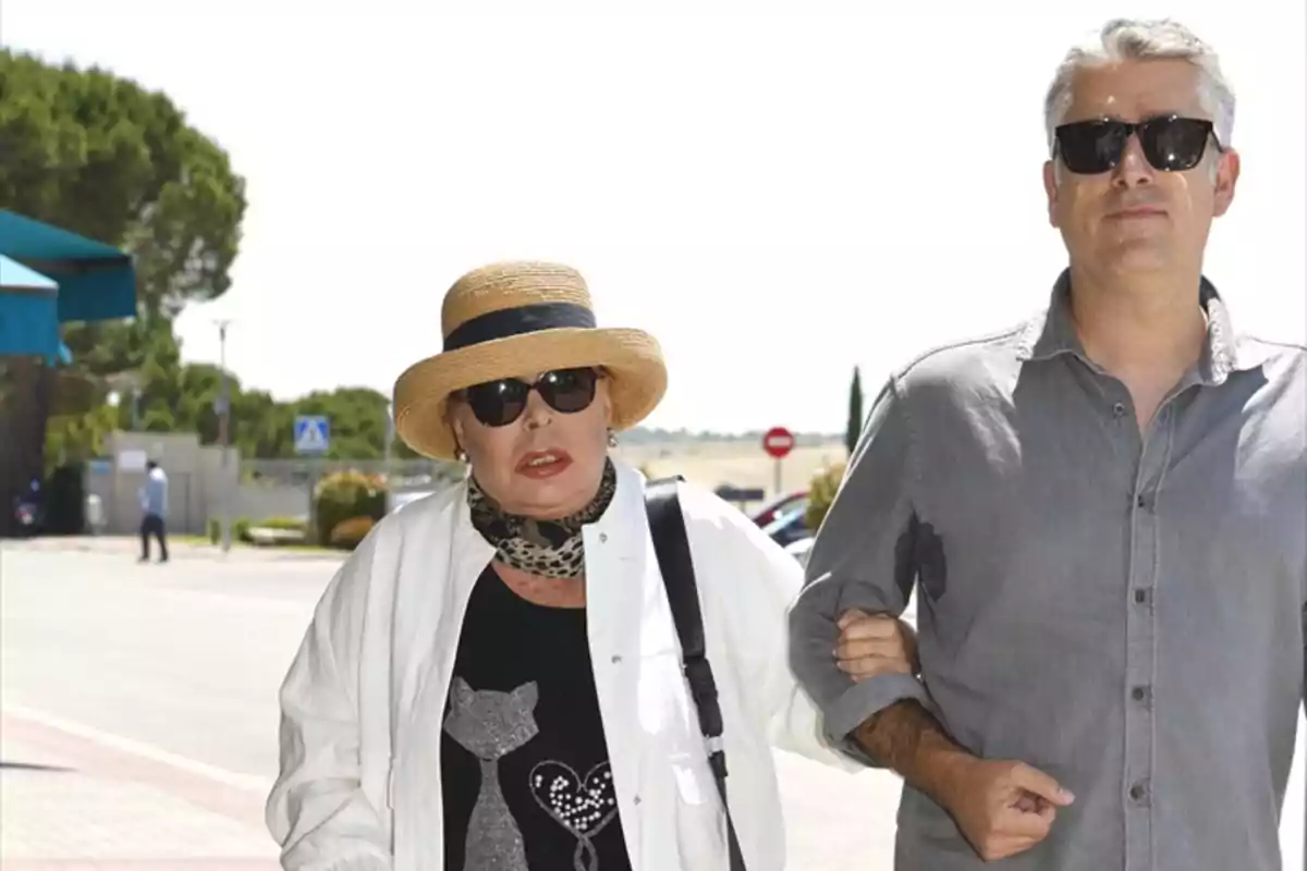Una mujer con sombrero y gafas de sol camina del brazo de un hombre con camisa gris en un entorno al aire libre. Una mujer con sombrero y gafas de sol camina del brazo de un hombre con camisa gris en un entorno al aire libre.