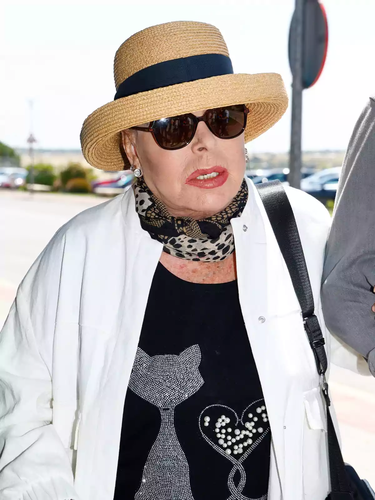 Older woman wearing a straw hat, sunglasses, polka dot scarf, and a black t-jersey with a silver cat design, walking outdoors on a sunny day Older woman wearing a straw hat, sunglasses, polka dot scarf, and a black t-jersey with a silver cat design, walking outdoors on a sunny day