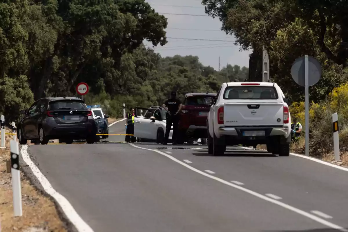 Una carretera está bloqueada por varios vehículos, incluyendo coches de policía, mientras agentes de seguridad inspeccionan la escena.
