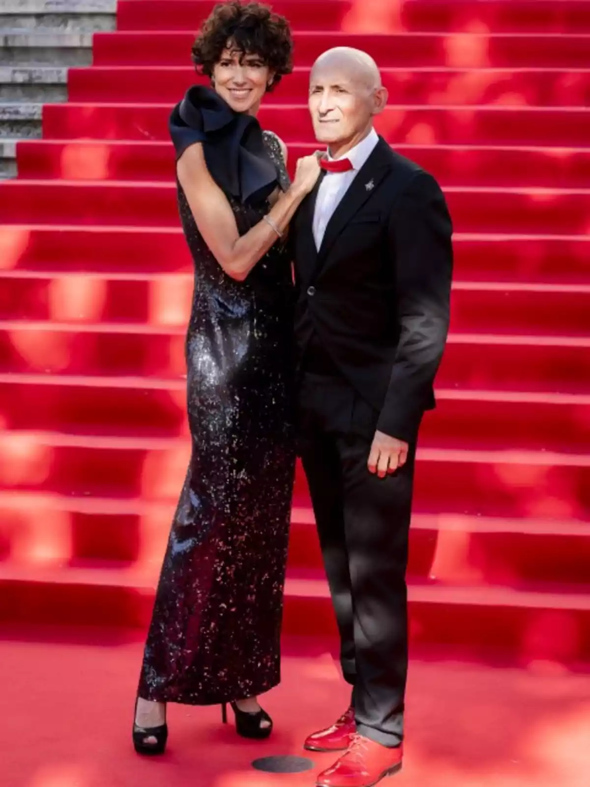 Una pareja elegantemente vestida posa en una alfombra roja con escaleras de fondo.