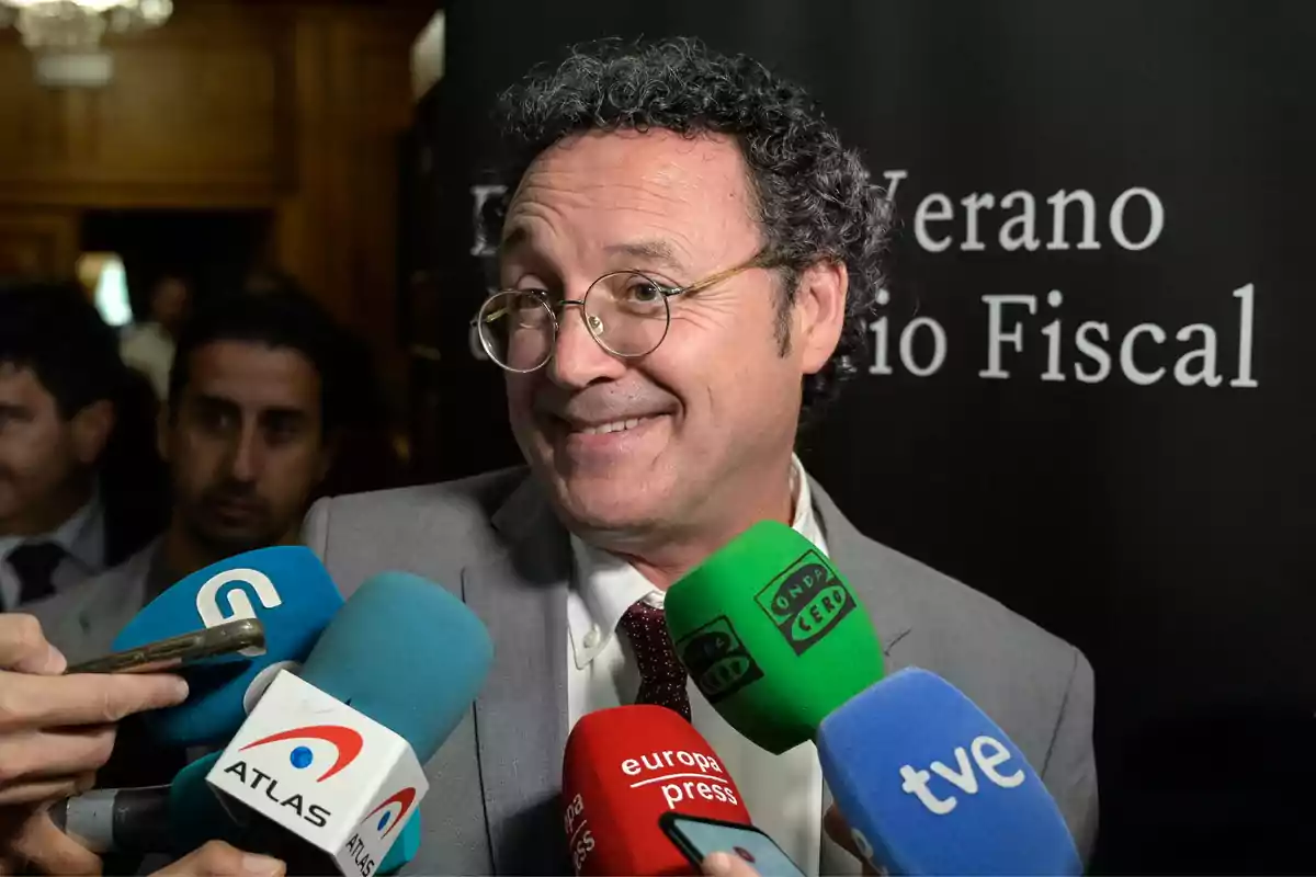 Un hombre con gafas y traje sonríe mientras es entrevistado por varios medios de comunicación que sostienen micrófonos de diferentes colores.