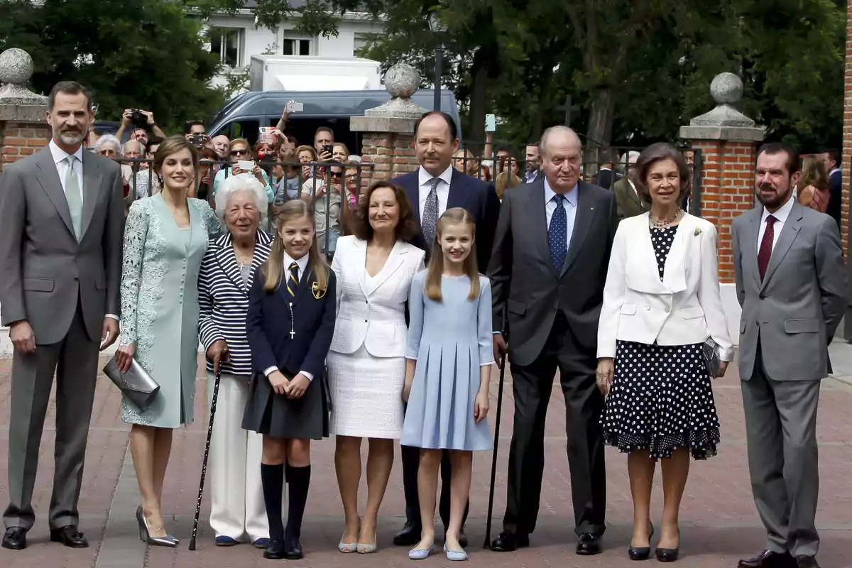 Un grupo de personas elegantemente vestidas posa para una foto al aire libre frente a una entrada de ladrillo, con varias personas al fondo tomando fotos. Un grupo de personas elegantemente vestidas posa para una foto al aire libre frente a una entrada de ladrillo, con varias personas al fondo tomando fotos.