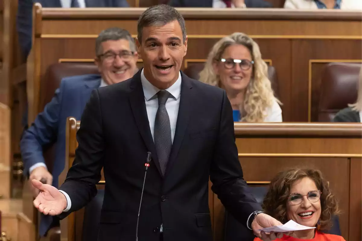 Un hombre de traje oscuro hablando en un podio con personas sonriendo en el fondo.