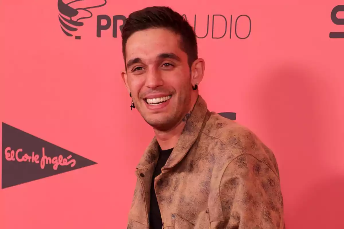 Un hombre sonriente con chaqueta marrón posando frente a un fondo rojo con logotipos.