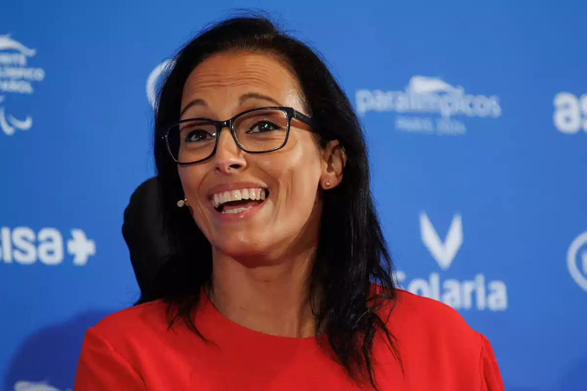 Mujer con gafas y camisa roja sonriendo frente a un fondo azul con logotipos.