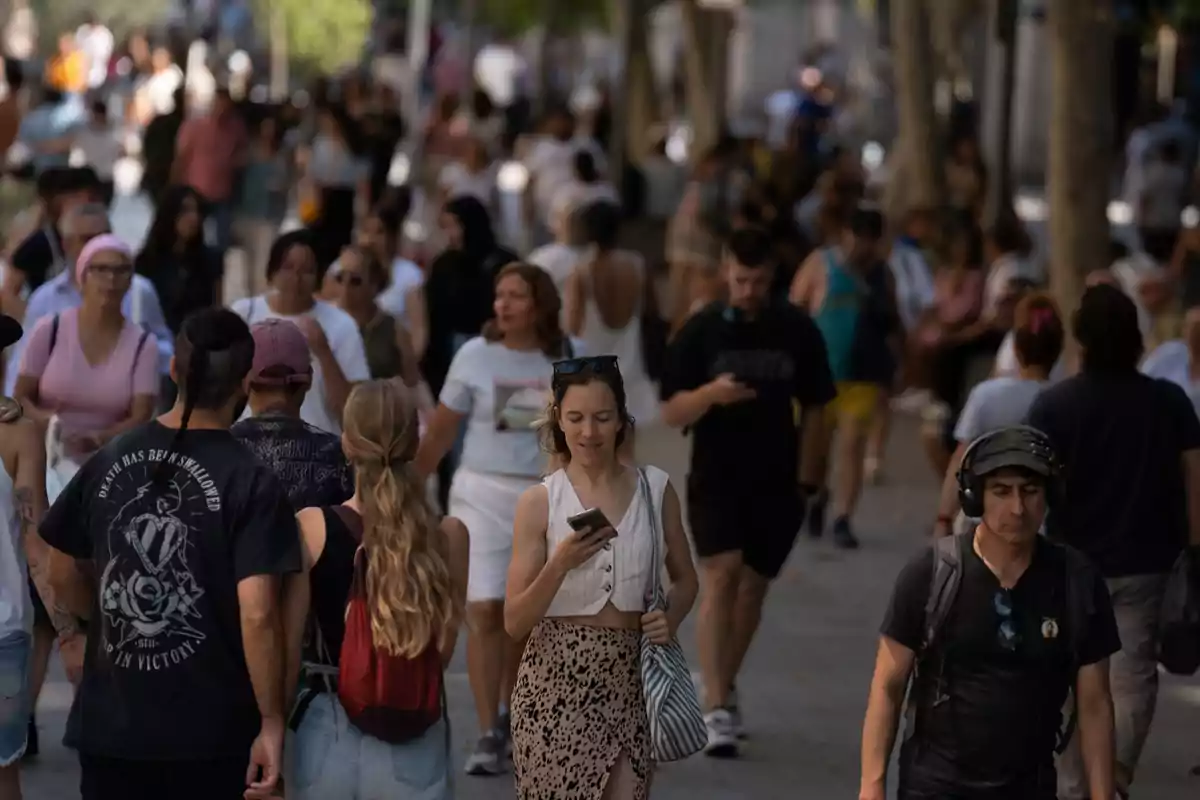 Una multitud de personas camina por una calle concurrida, algunas mirando sus teléfonos mientras otras conversan o disfrutan del paseo.