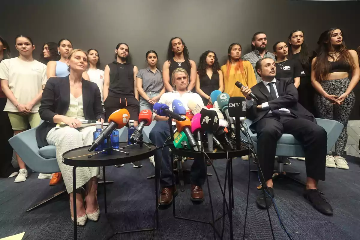 Una conferencia de prensa con varias personas sentadas frente a micrófonos y cámaras, mientras un grupo de personas está de pie detrás de ellos.