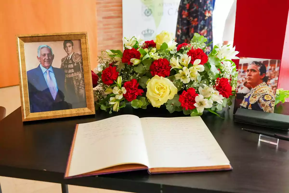 Una mesa con un libro abierto, un arreglo floral con flores rojas y blancas, y dos fotografías enmarcadas de personas.