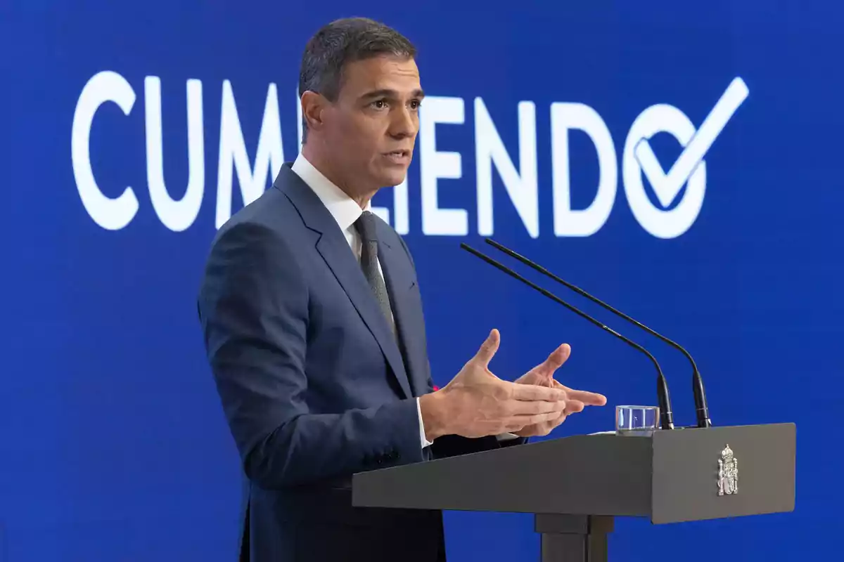 Un hombre en traje azul hablando en un podio con la palabra "CUMPLIENDO" en el fondo.