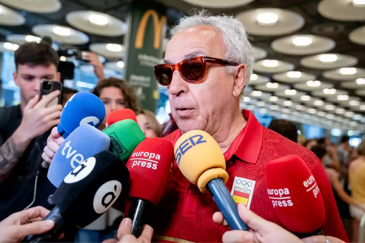 Un hombre con gafas de sol y una camiseta roja es entrevistado por varios medios de comunicación, rodeado de micrófonos de diferentes colores y logotipos.