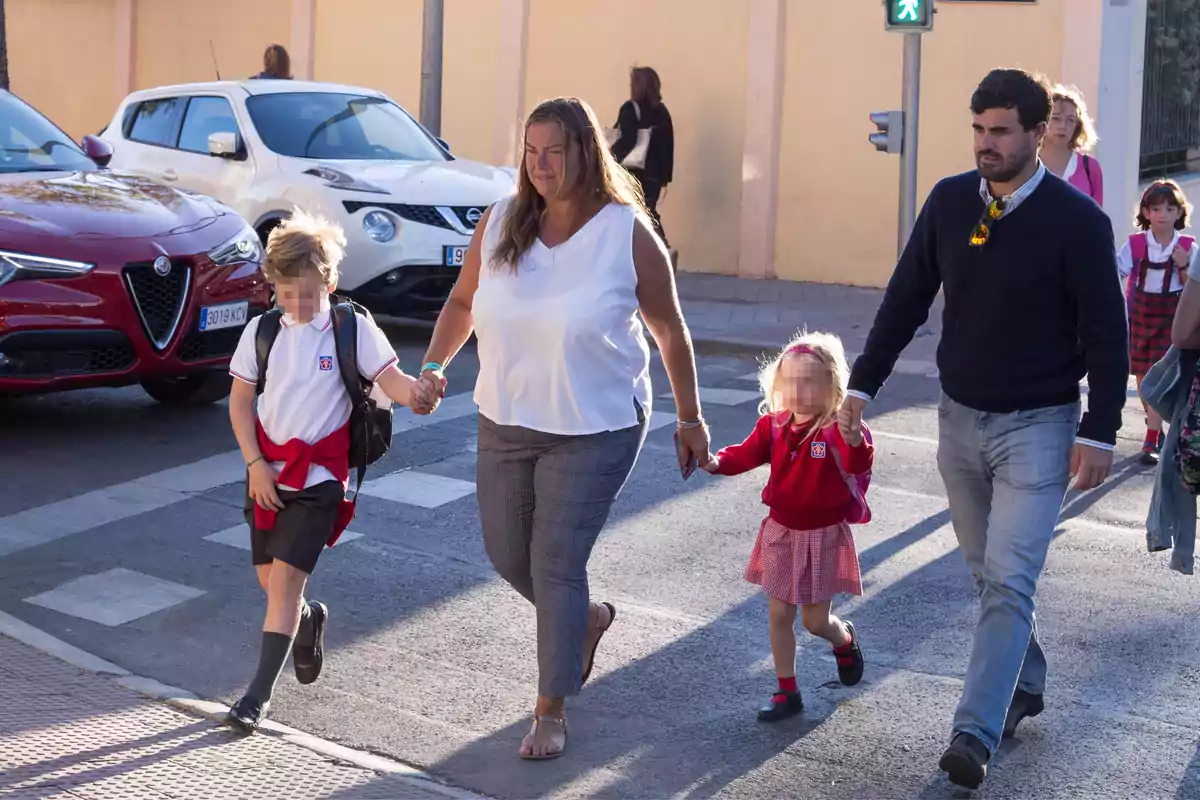 Una familia cruzando la calle con dos niños en uniforme escolar, acompañados por dos adultos, con coches estacionados al fondo.