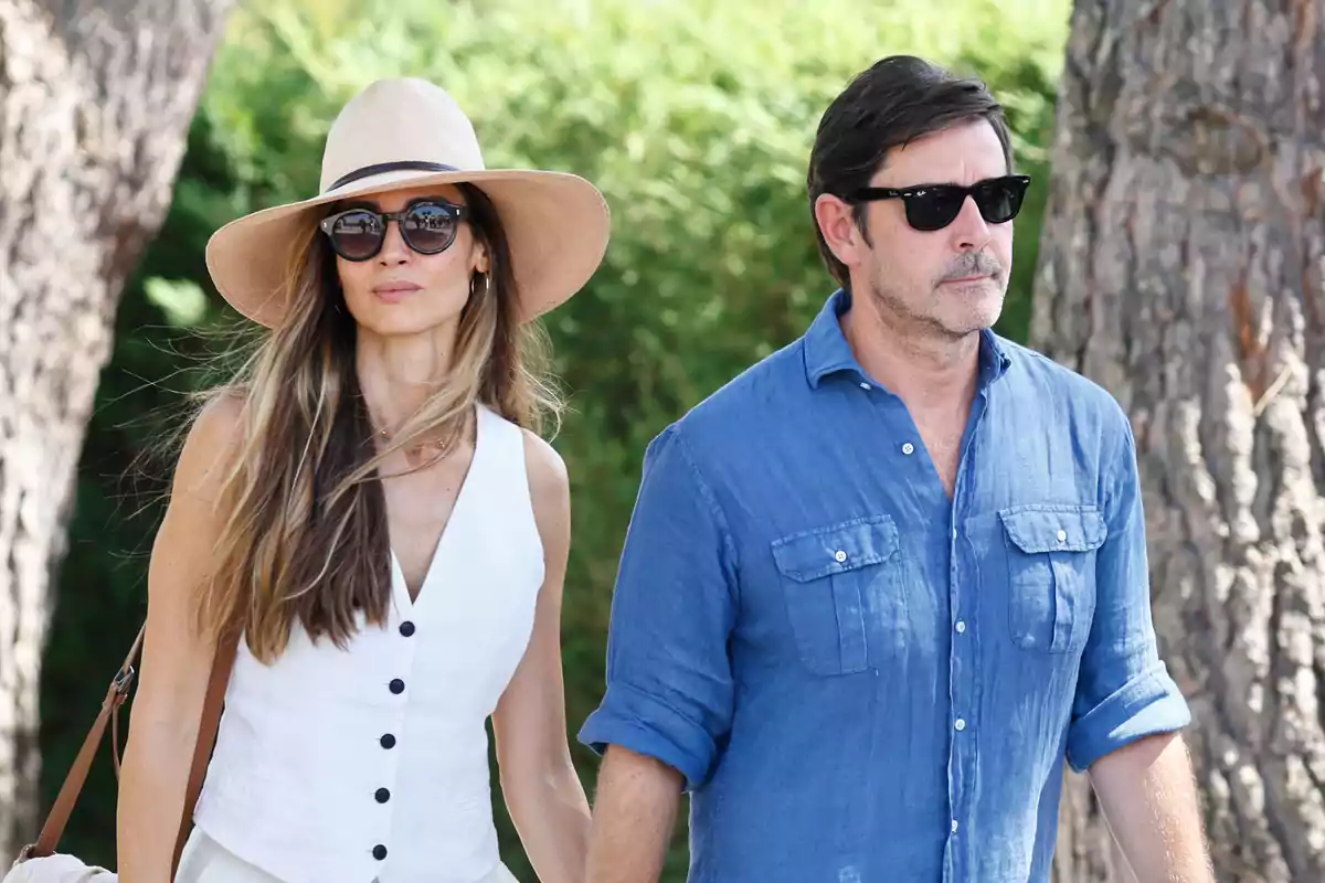 Una mujer con sombrero y gafas de sol camina junto a un hombre con camisa azul y gafas de sol en un entorno al aire libre con árboles.