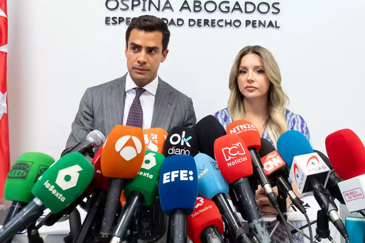 Dos personas en una conferencia de prensa rodeadas de micrófonos de diferentes medios de comunicación.