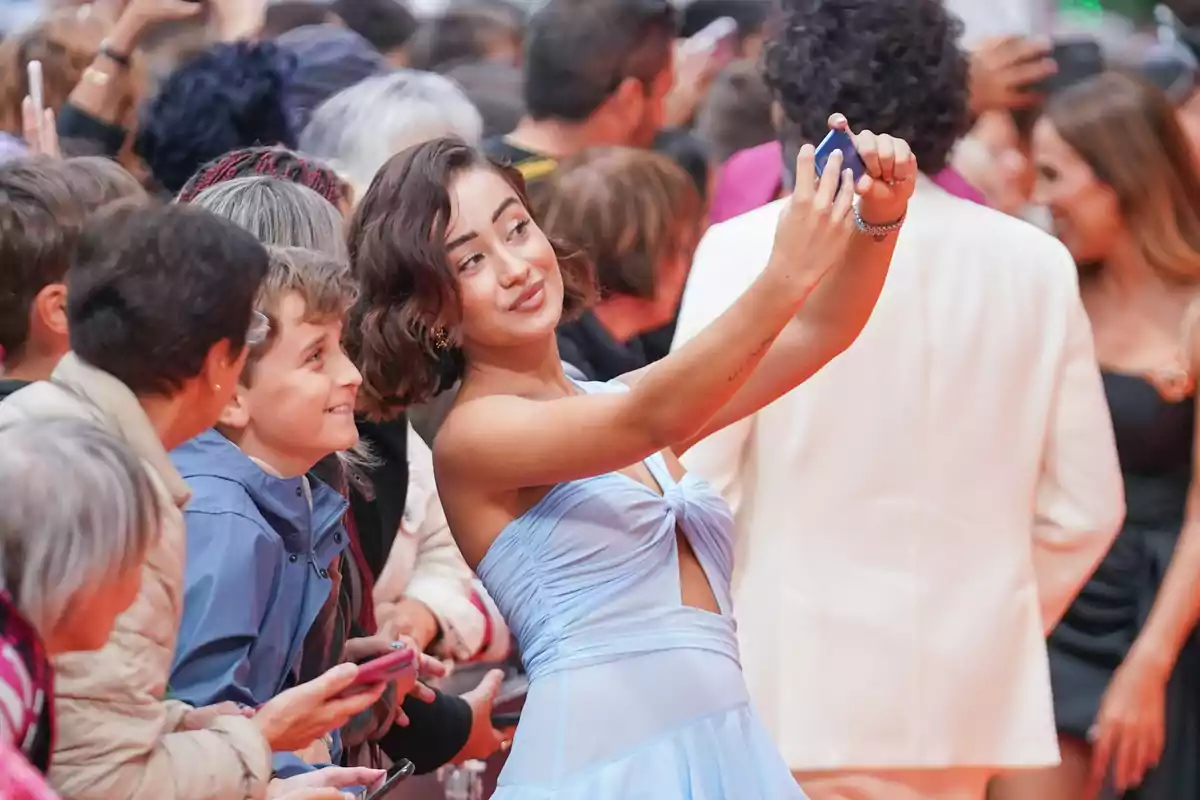 Una mujer con un vestido azul claro se toma una selfie con un grupo de personas en un evento concurrido.