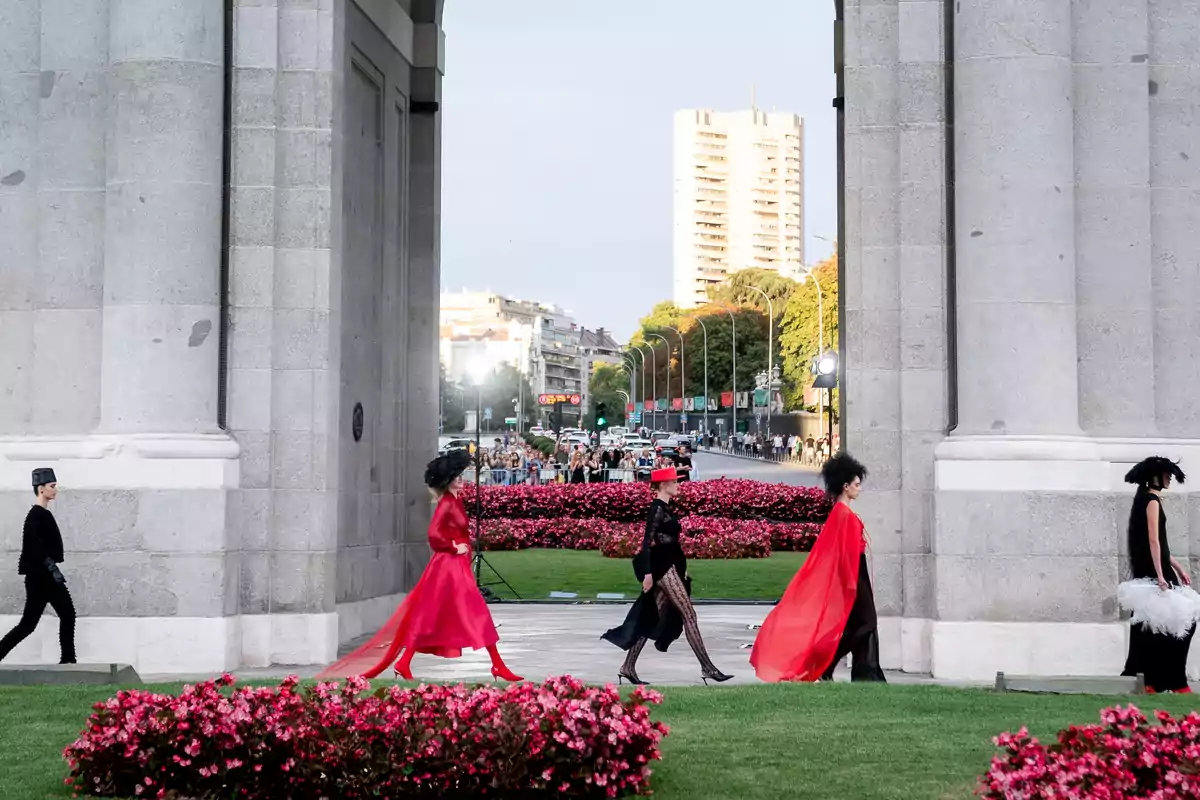 Desfile de moda al aire libre con modelos caminando entre columnas de piedra, rodeadas de flores y con un edificio alto al fondo.