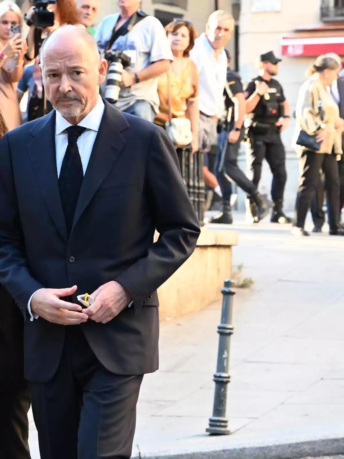 Hombre calvo con barba vestido con traje oscuro y corbata negra caminando por la calle mientras varias personas y fotógrafos están al fondo Hombre calvo con barba vestido con traje oscuro y corbata negra caminando por la calle mientras varias personas y fotógrafos están al fondo