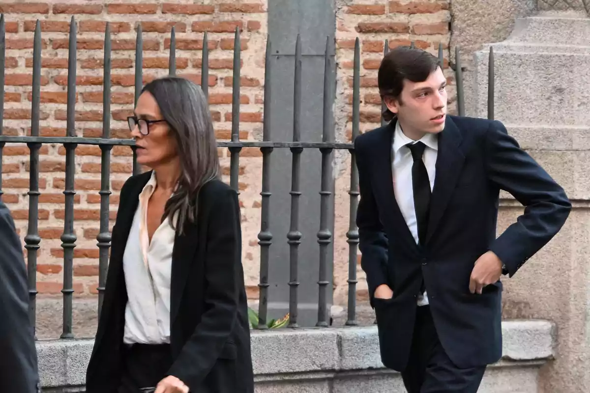 Dos personas vestidas de manera formal caminan por una calle con una pared de ladrillos y una reja de hierro detrás de ellas. Dos personas vestidas de manera formal caminan por una calle con una pared de ladrillos y una reja de hierro detrás de ellas.