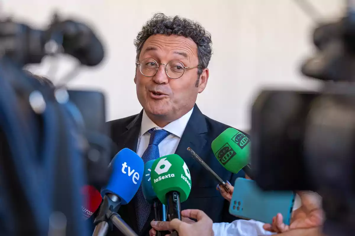 Un hombre con gafas y traje oscuro habla frente a varios micrófonos de diferentes medios de comunicación.