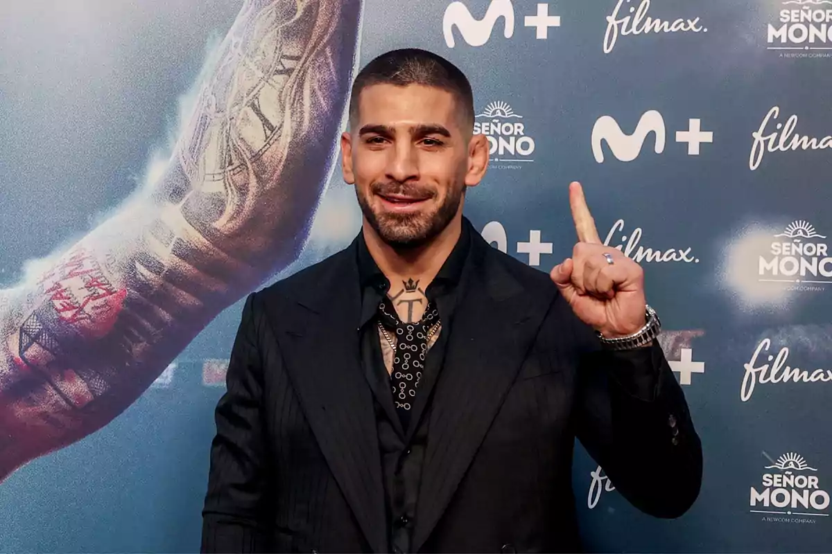 Hombre con traje oscuro y tatuajes en el cuello y brazo, posando frente a un fondo con logotipos de "Movistar+", "Filmax" y "Señor Mono", levantando el dedo índice de su mano derecha.