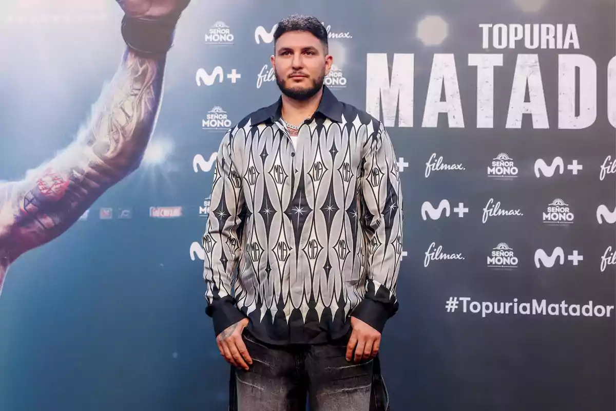 Un hombre con barba y camisa de diseño geométrico posando frente a un fondo promocional que incluye los logotipos de Movistar Plus, Filmax y Señor Mono, así como el hashtag #TopuriaMatador.