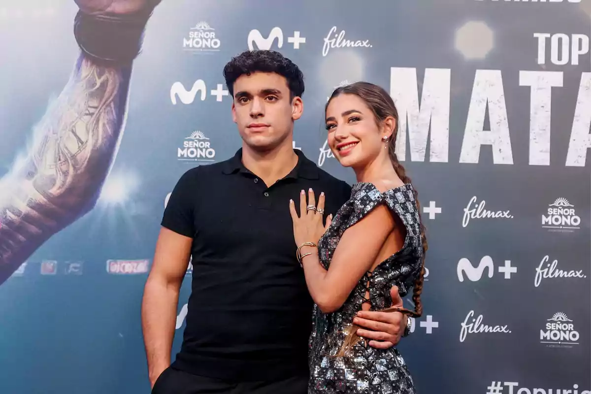 Dos personas posando en un evento con un fondo promocional de una película o serie.