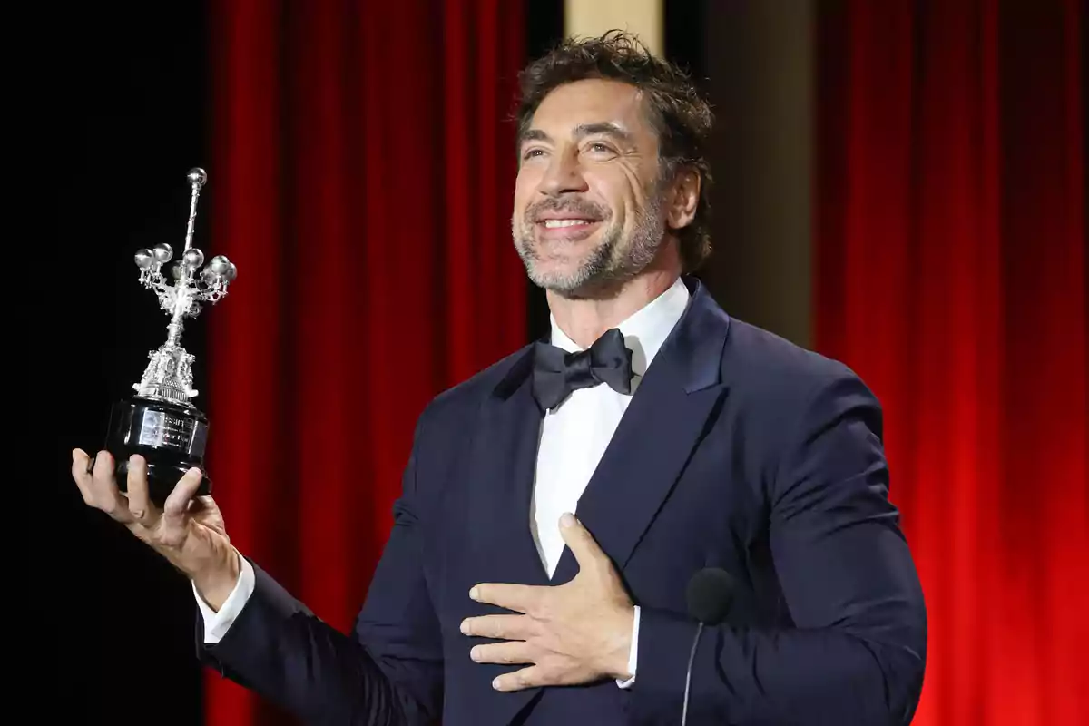 Hombre con traje y pajarita sosteniendo un trofeo en un escenario con cortinas rojas.