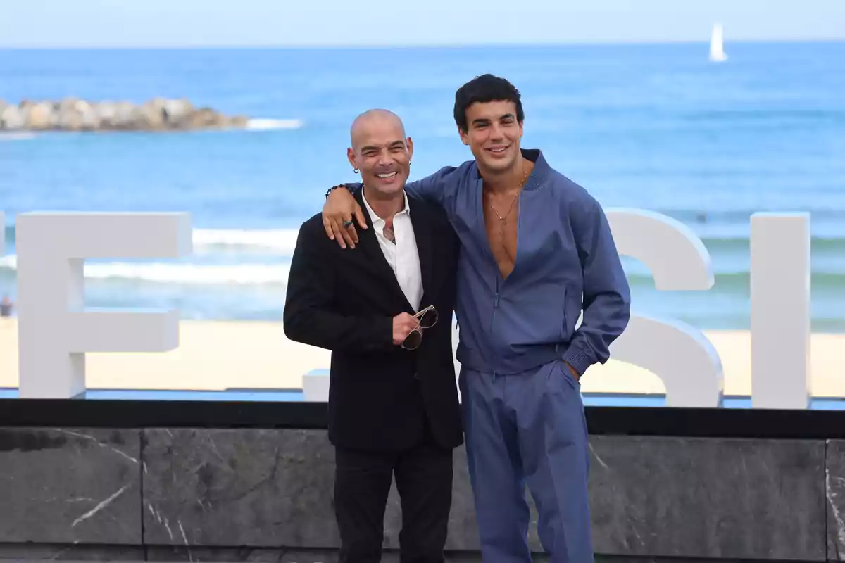 Dos personas posan sonrientes frente al mar con letras grandes de fondo.