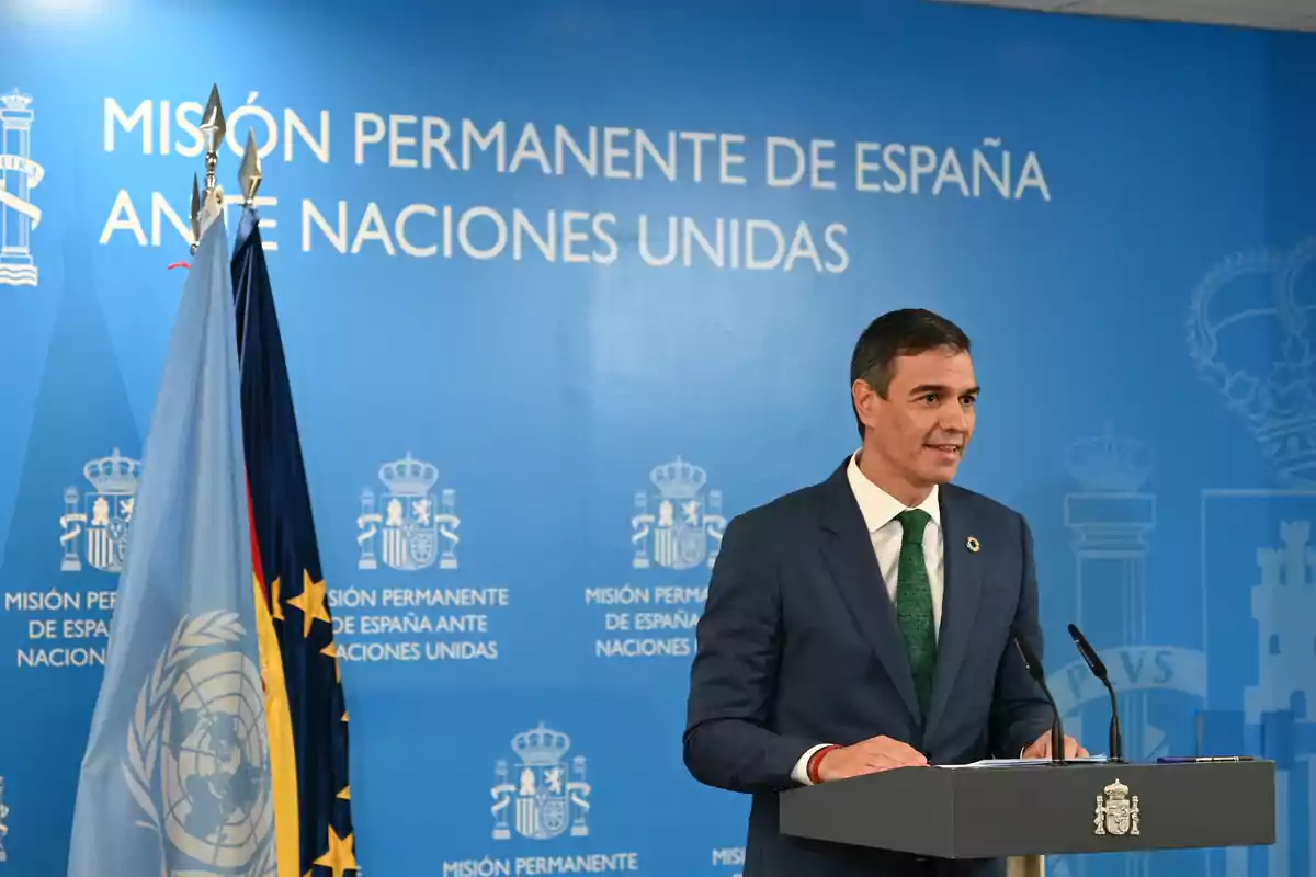 Un hombre en traje da un discurso en un podio frente a un fondo azul que dice "Misión Permanente de España ante Naciones Unidas", con banderas de la ONU y de España a su lado.