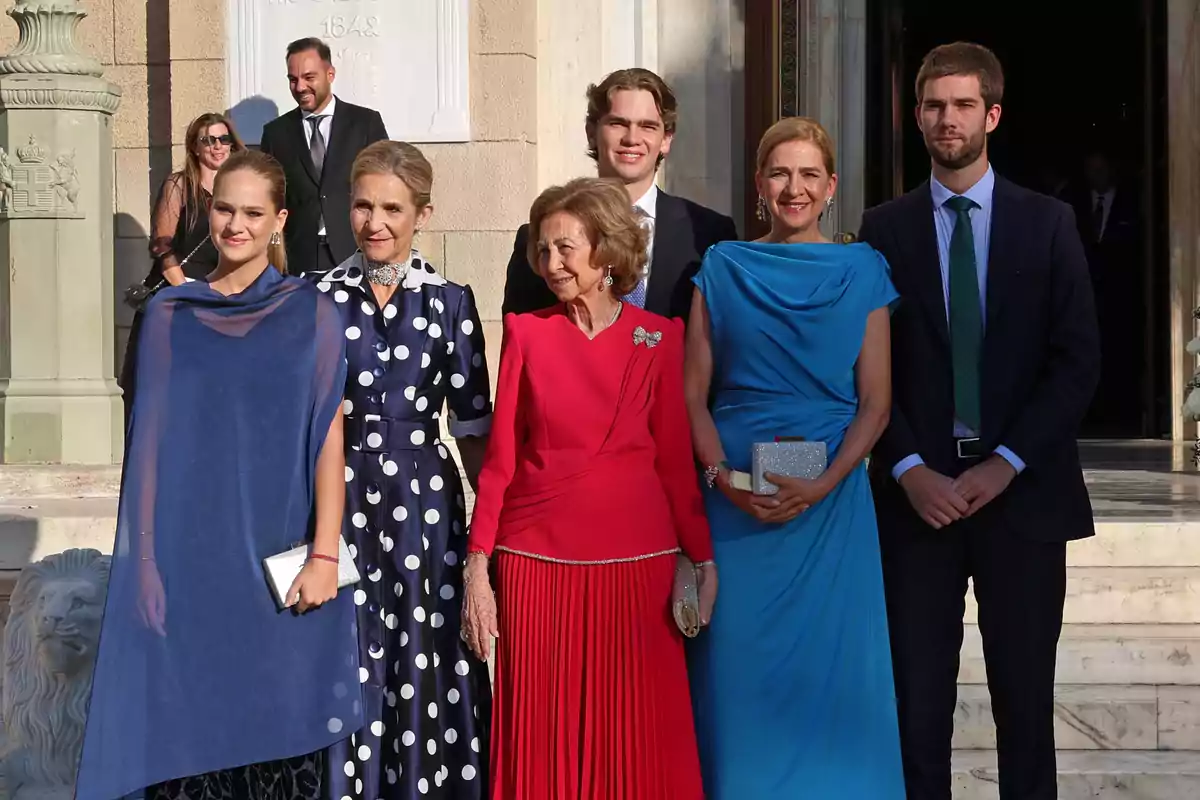 Grupo de seis personas elegantemente vestidas posando en la entrada de un edificio, tres mujeres llevan vestidos largos en tonos azul y rojo, dos hombres usan traje y corbata, todos sonríen y parecen estar en un evento formal Grupo de seis personas elegantemente vestidas posando en la entrada de un edificio, tres mujeres llevan vestidos largos en tonos azul y rojo, dos hombres usan traje y corbata, todos sonríen y parecen estar en un evento formal