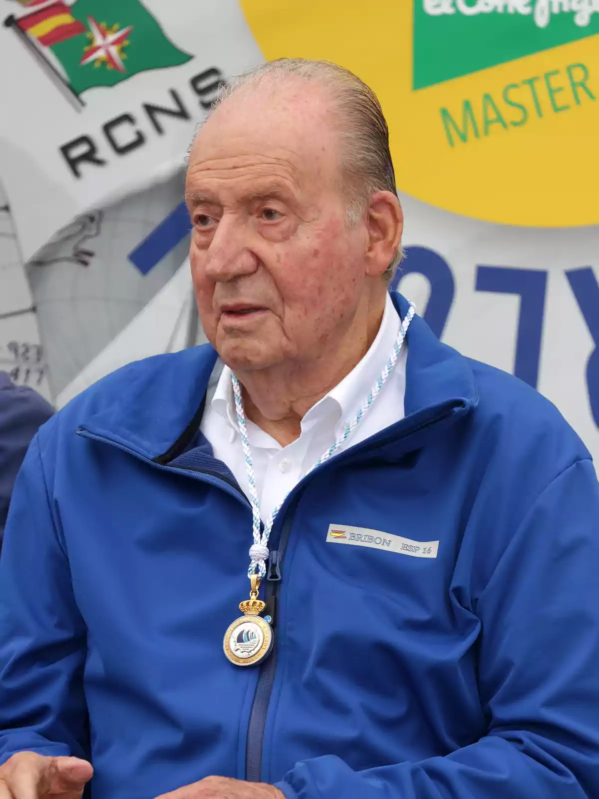 Un hombre mayor con chaqueta azul y medalla, frente a un fondo con logotipos y texto. Un hombre mayor con chaqueta azul y medalla, frente a un fondo con logotipos y texto.