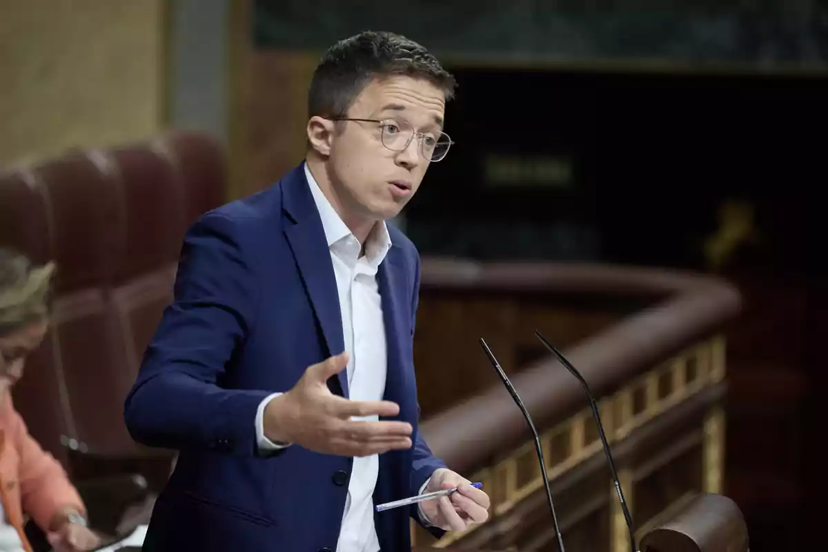 Un hombre con gafas y traje azul habla gesticulando en un podio en un entorno formal.