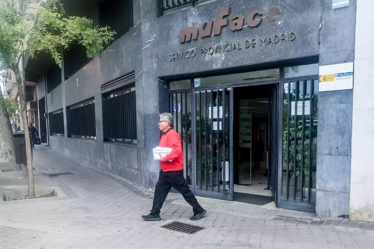 Un hombre con suéter rojo camina frente a un edificio gris con el letrero 