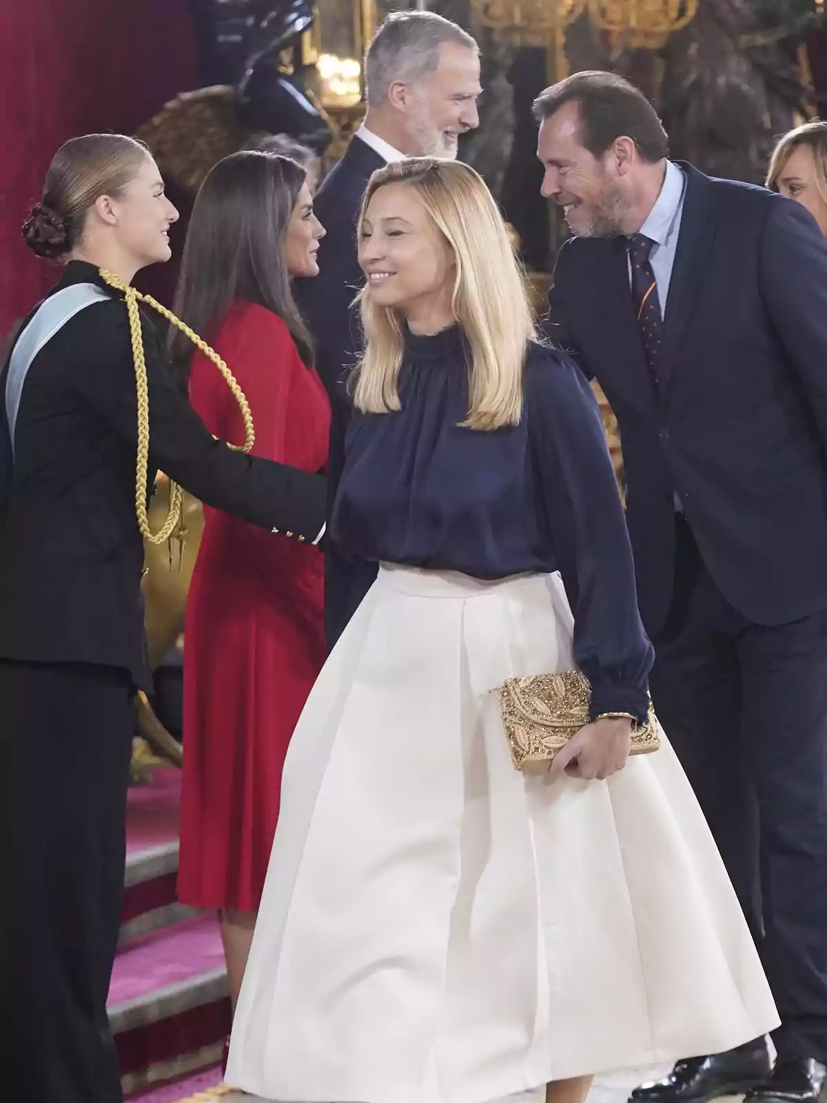 Un grupo de personas elegantemente vestidas se saludan y conversan en un evento formal en un entorno decorado con detalles dorados y rojos. Un grupo de personas elegantemente vestidas se saludan y conversan en un evento formal en un entorno decorado con detalles dorados y rojos.