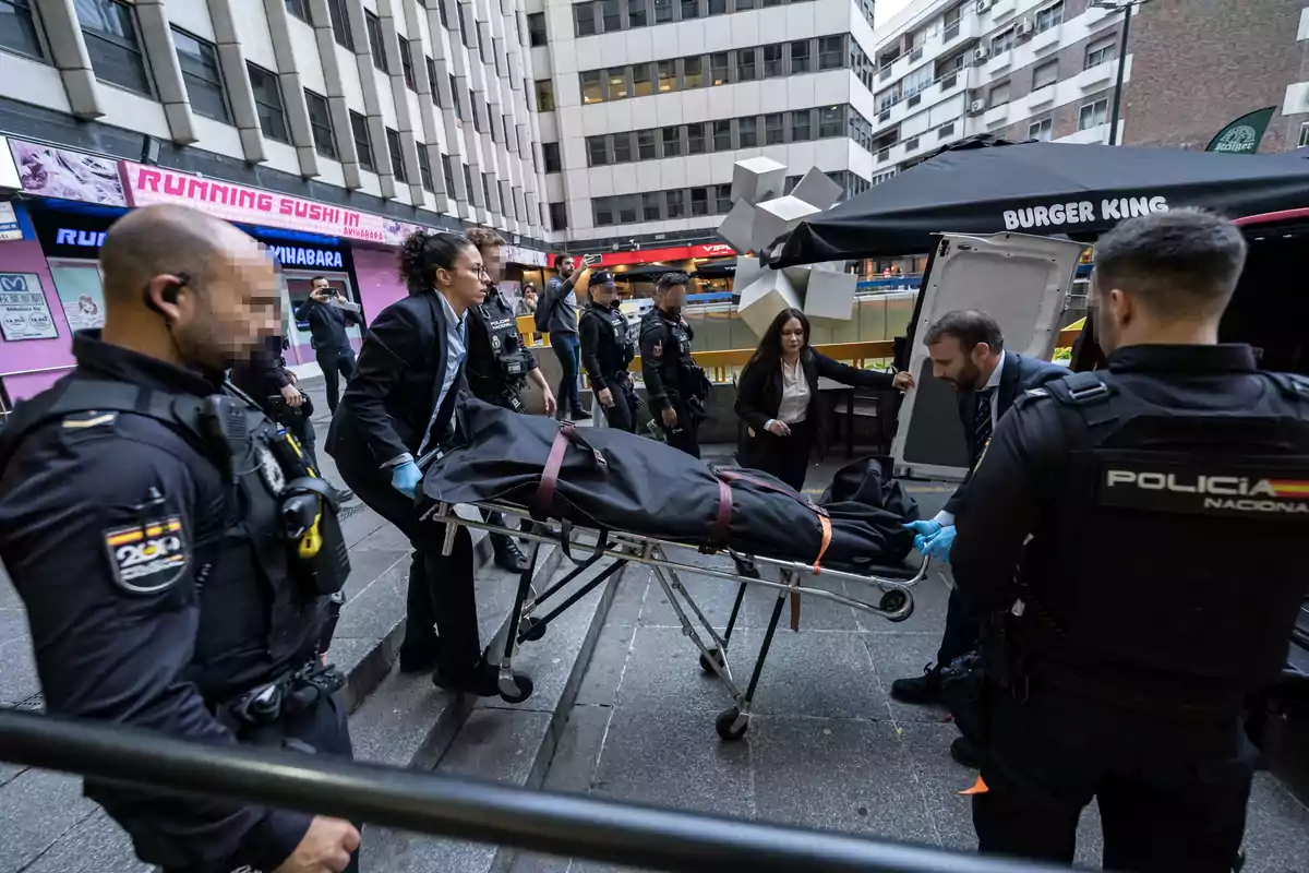 Un grupo de personas, incluyendo agentes de policía, transporta una camilla con un cuerpo cubierto en una calle urbana.