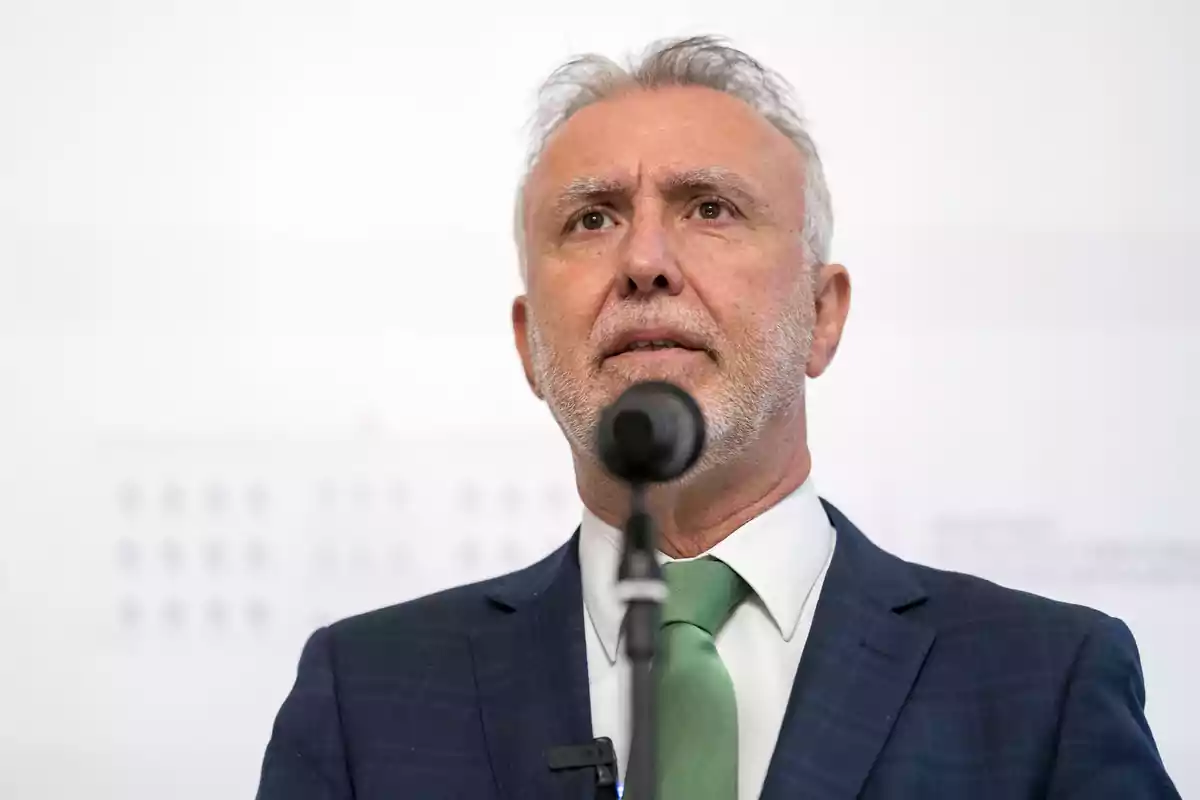 Hombre de cabello canoso y barba hablando frente a un micrófono con traje azul y corbata verde.