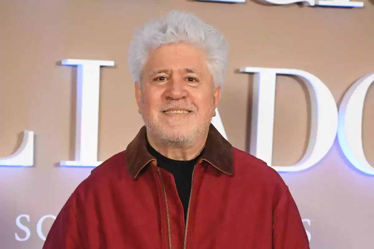 Un hombre con cabello canoso y barba ligera viste una chaqueta roja frente a un fondo con letras grandes.