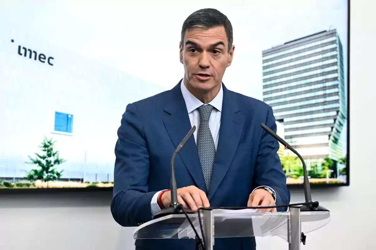 Un hombre en traje azul hablando en un podio con micrófonos frente a un edificio moderno.