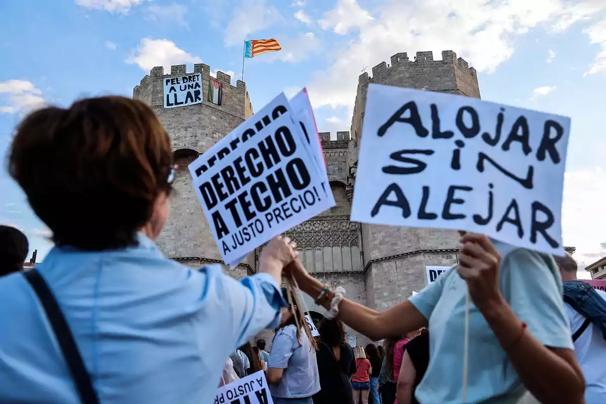 Personas sostienen pancartas en una manifestación frente a una torre histórica, con mensajes sobre el derecho a la vivienda.