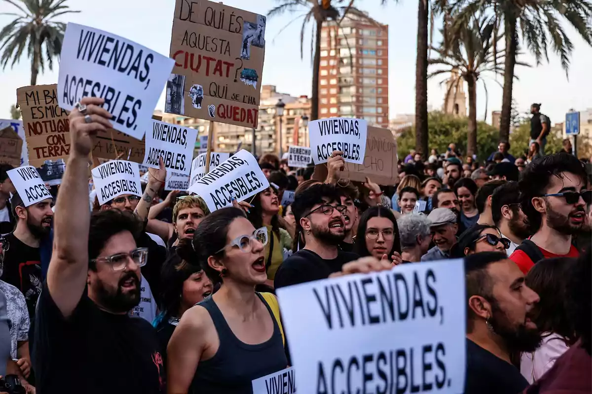 Un grupo de personas se manifiesta sosteniendo carteles que piden viviendas accesibles en un entorno urbano con palmeras y edificios al fondo.