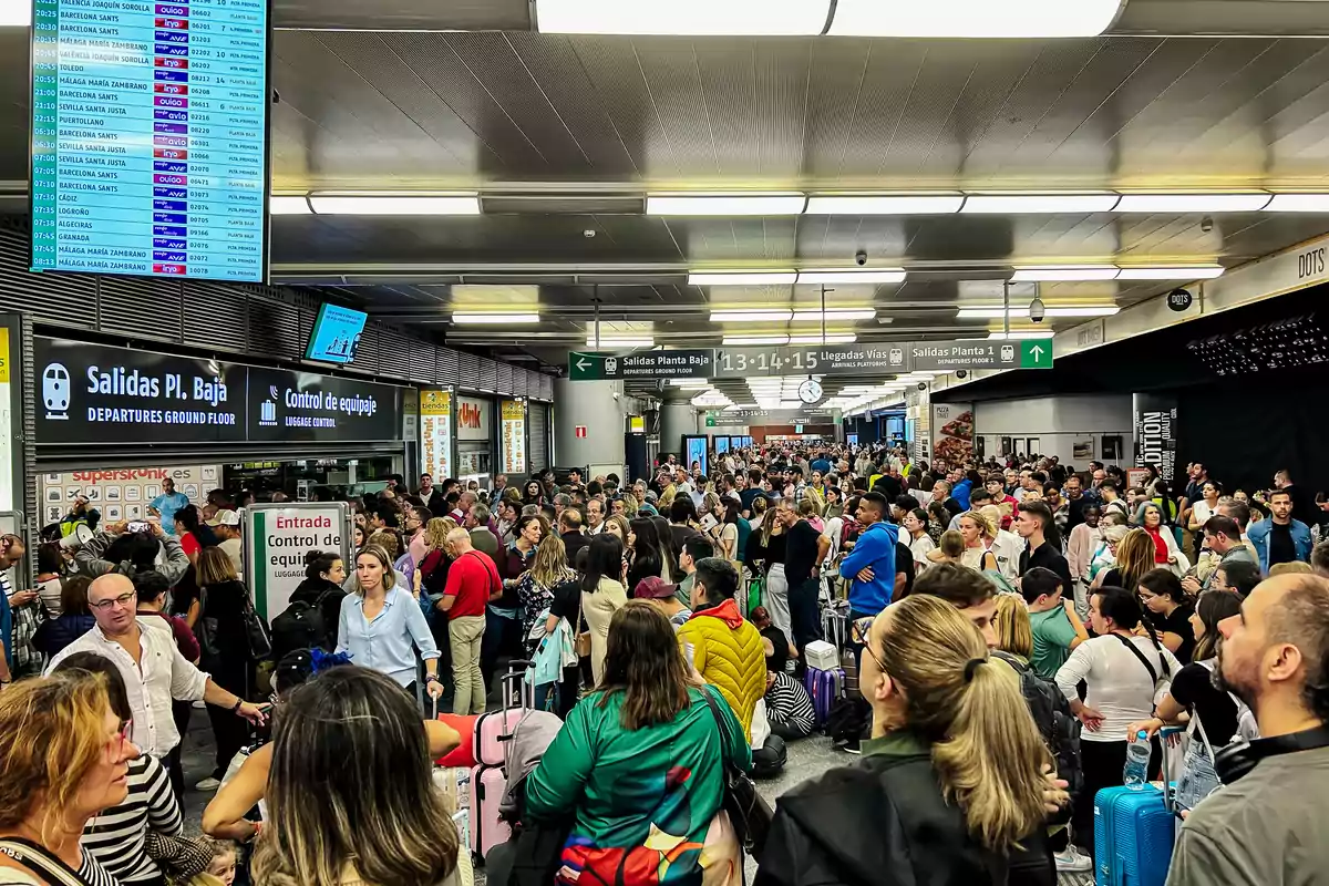 Una multitud de personas se aglomera en una estación de tren, con pantallas de información y señales de salidas y llegadas visibles en el techo.