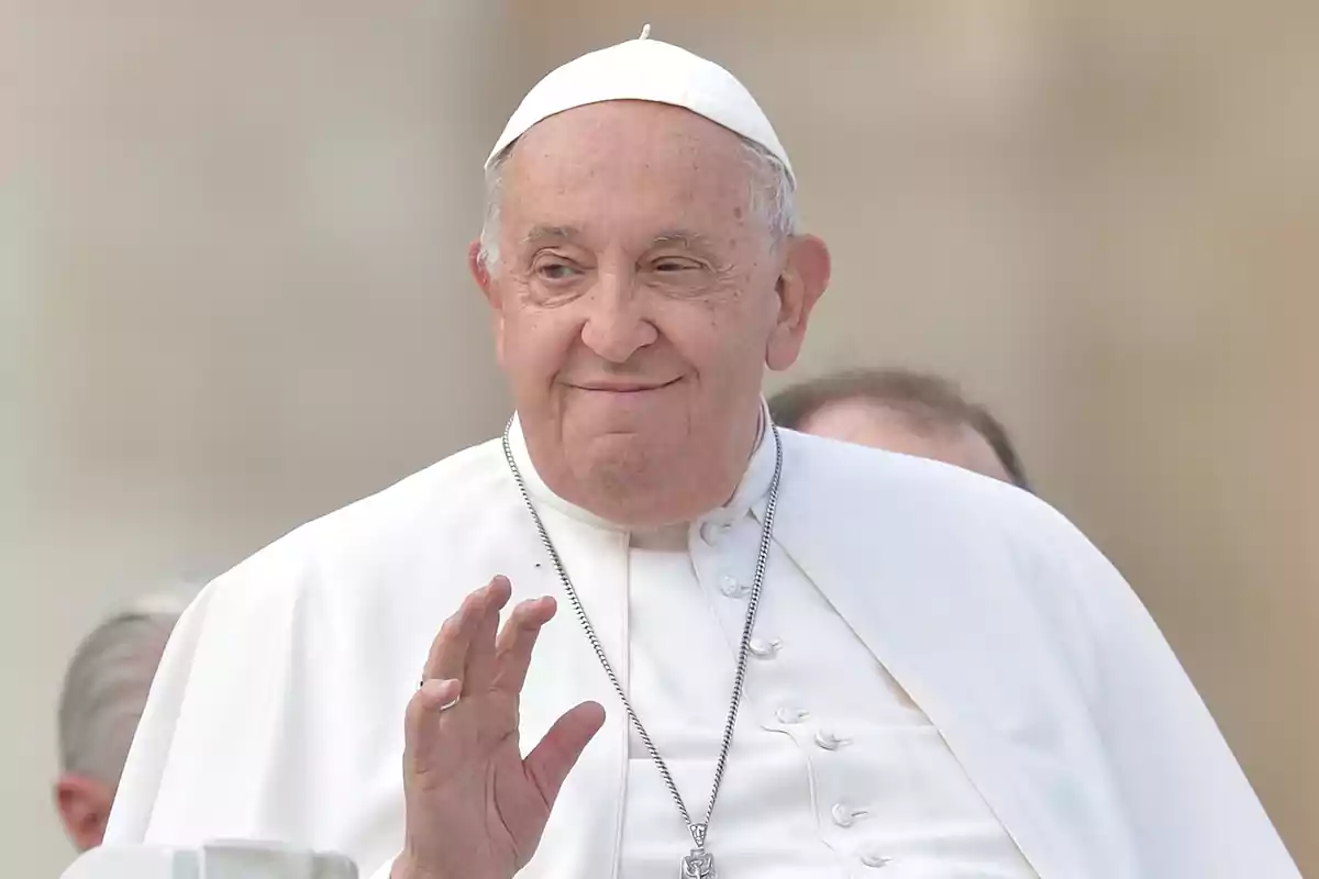 Un hombre mayor vestido con atuendo religioso blanco y un sombrero pequeño saluda con la mano.