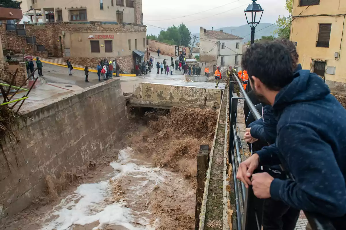 Personas observan un torrente de agua marrón que fluye con fuerza por un canal en una zona urbana mientras algunas personas se agrupan en la calle y otras miran desde un puente.