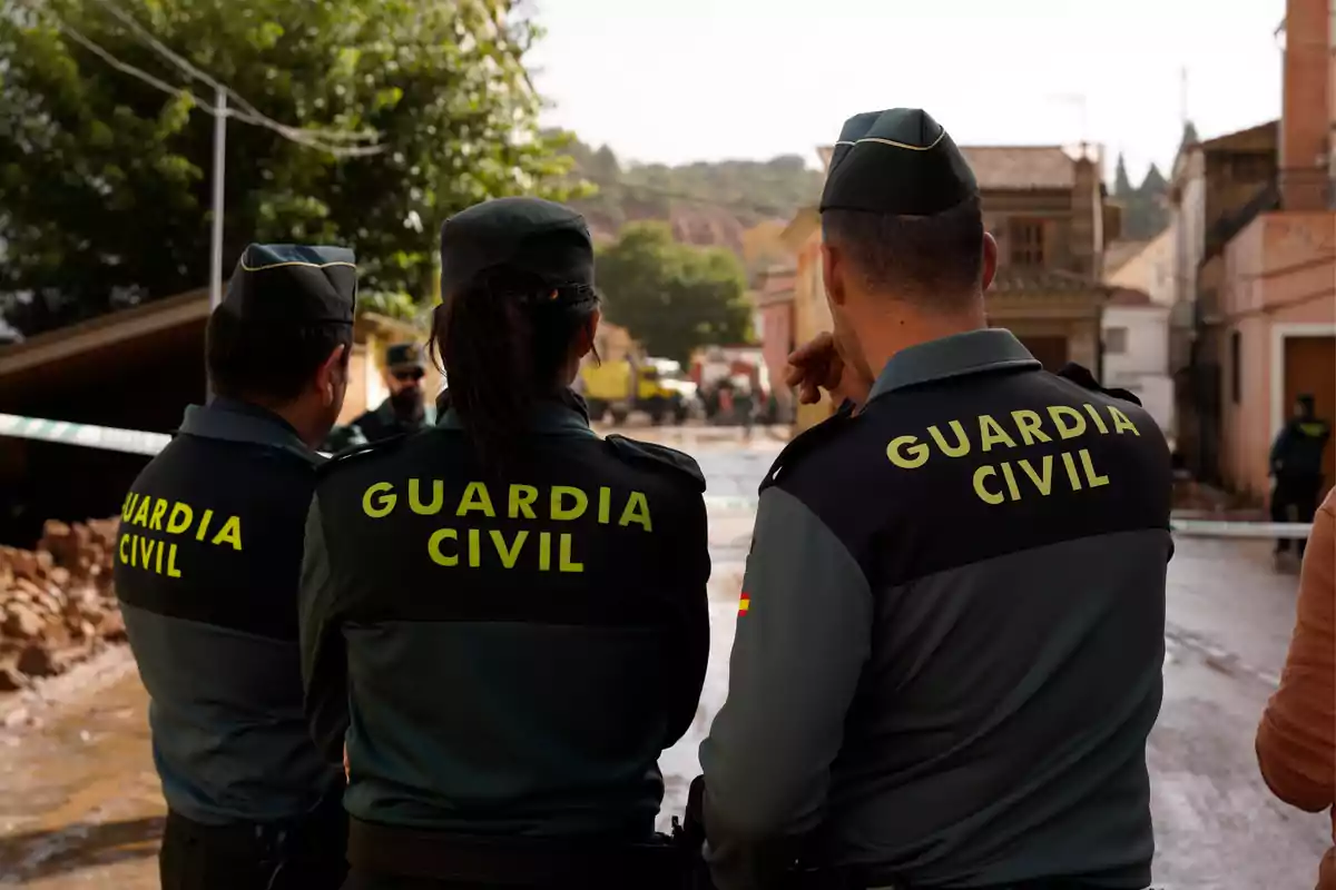 Tres agentes de la Guardia Civil de espaldas observan una escena en una calle con casas y árboles al fondo.