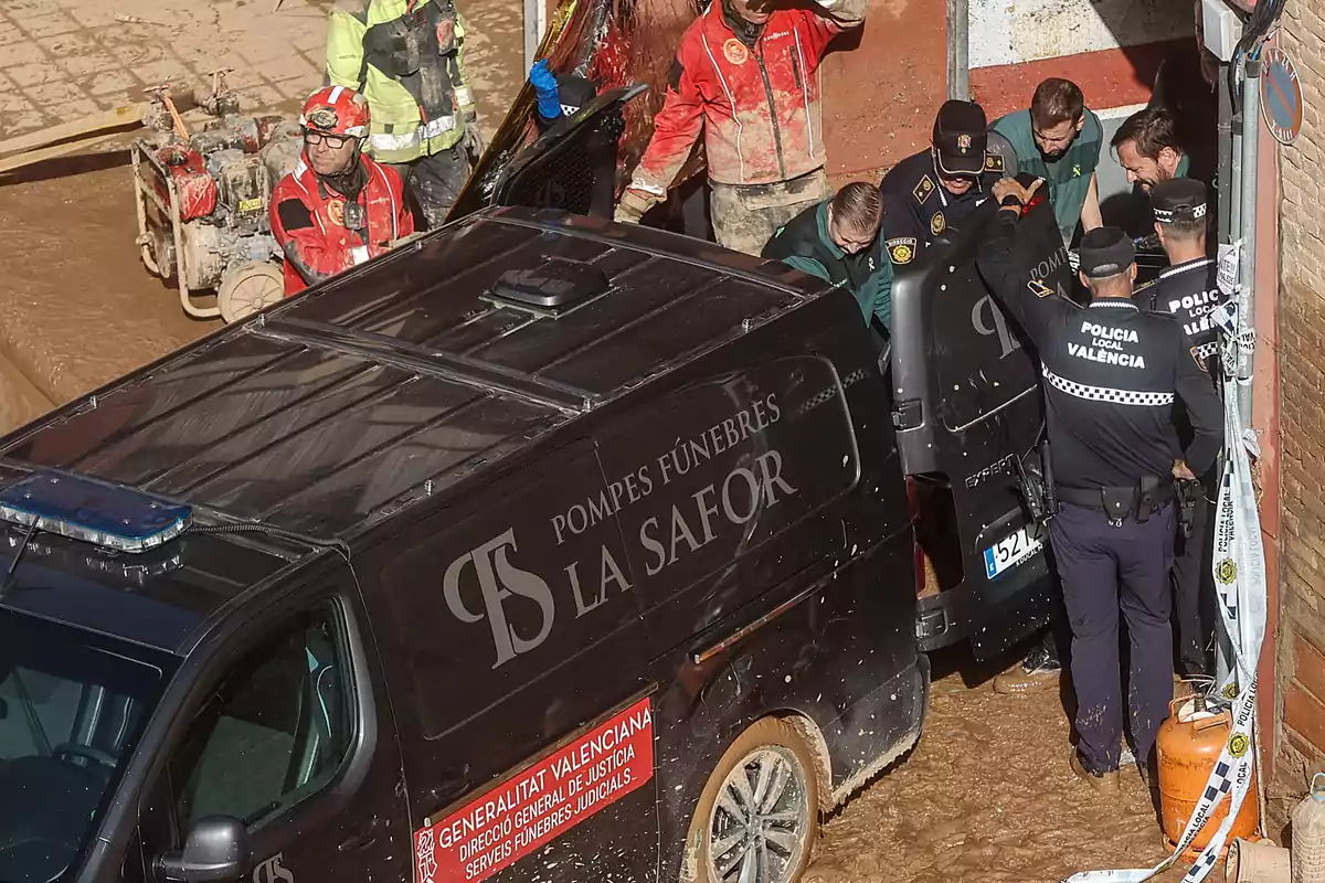 Un grupo de personas, incluyendo personal de emergencias y policía, se encuentra alrededor de un vehículo fúnebre en una zona con barro.