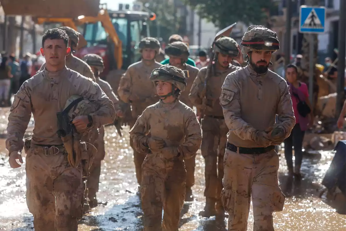 Un grupo de soldados camina por una calle llena de barro y agua, vistiendo uniformes y cascos, mientras realizan labores de ayuda en una zona afectada por un desastre natural.