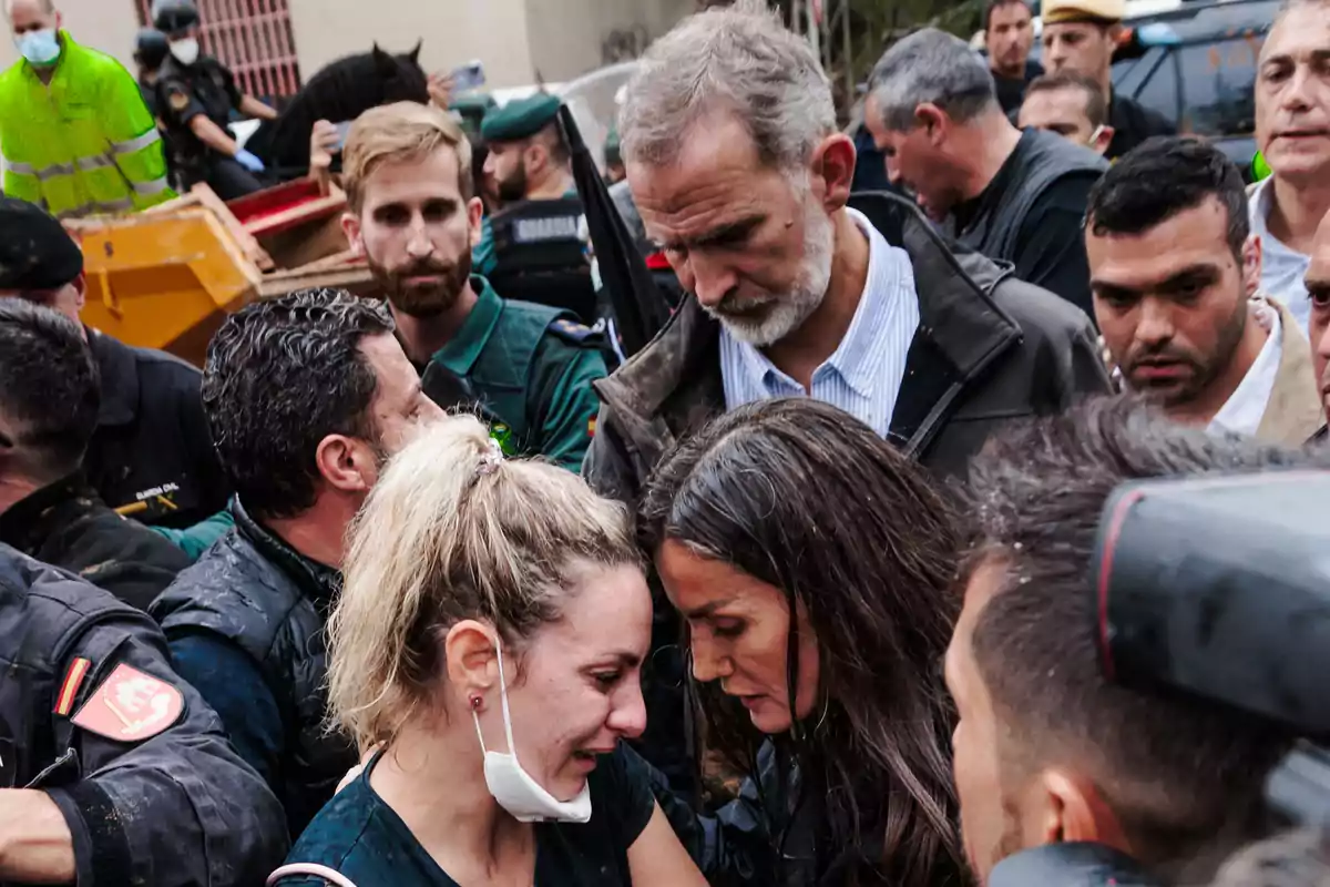 Un grupo de personas se reúne en un entorno al aire libre, algunas de ellas parecen emocionadas o preocupadas, mientras otras observan la escena.