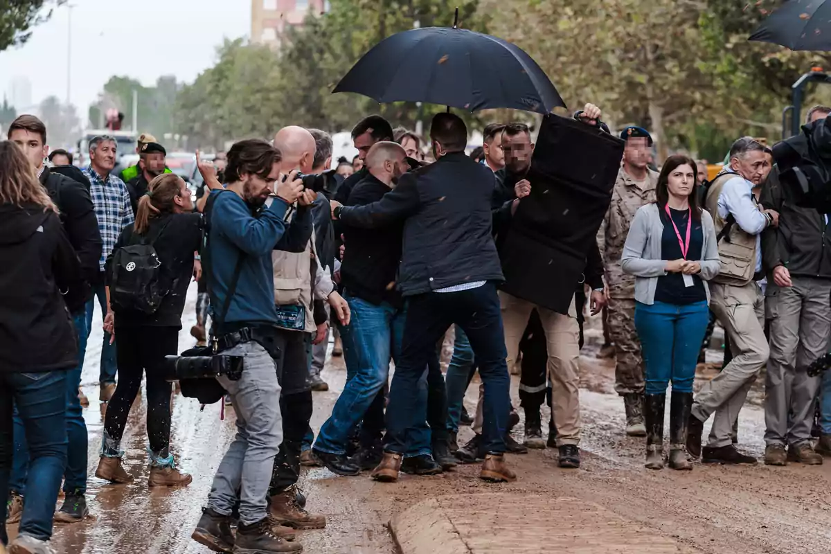 Un grupo de personas, algunas con cámaras y otras con paraguas, se encuentra en una calle embarrada rodeada de árboles.